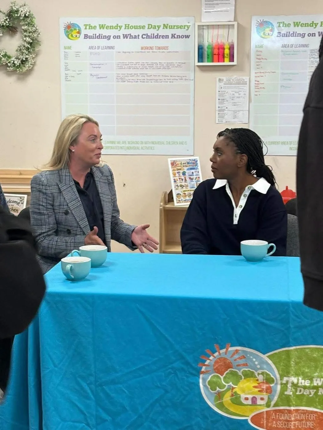 Victoria Vann and Kemi Badenoch talking across a table with a blue Wendy House Day Nursery cloth, nursery planning boards on the wall behind